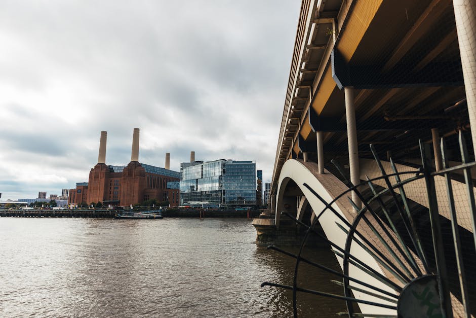 A view of the River Thames from the riverside with a large modern building and the historic Battersea Power Station with four tall chimneys in the background, under an overcast sky. In the foreground,