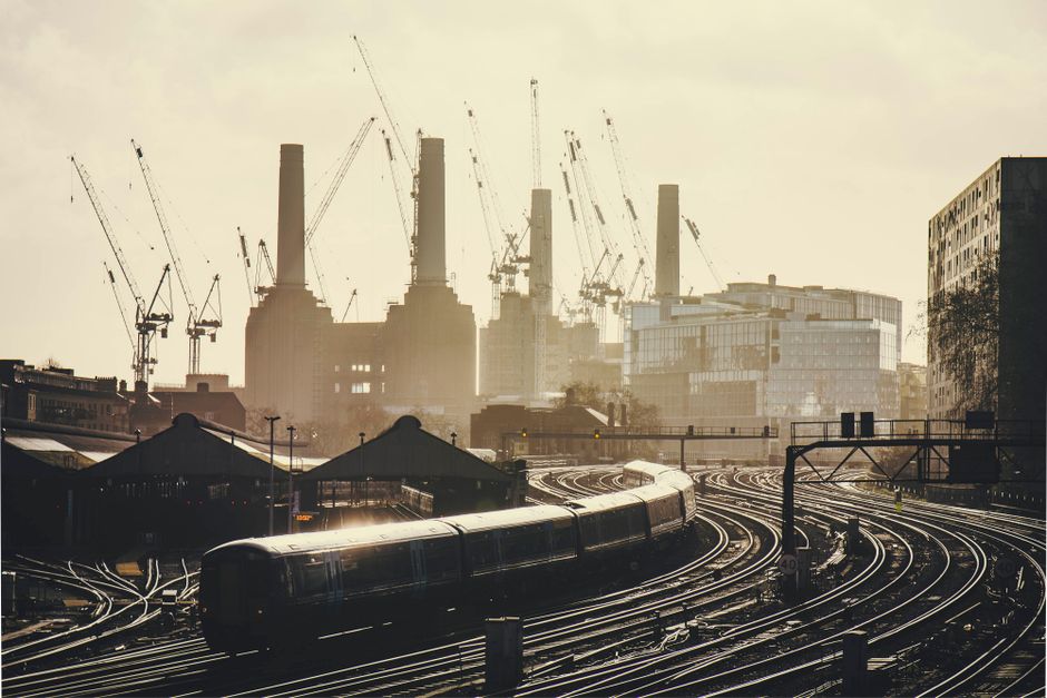 A view of the River Thames from the riverside with a large modern building and the historic Battersea Power Station with four tall chimneys in the background, under an overcast sky. In the foreground,