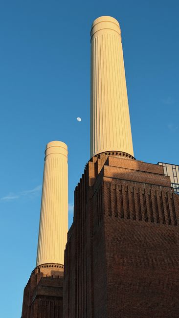 A large industrial building with three tall smokestacks, made of red brick, situated on the riverbank near central London. To the right, a modern glass office building reflects the cloudy sky, contras