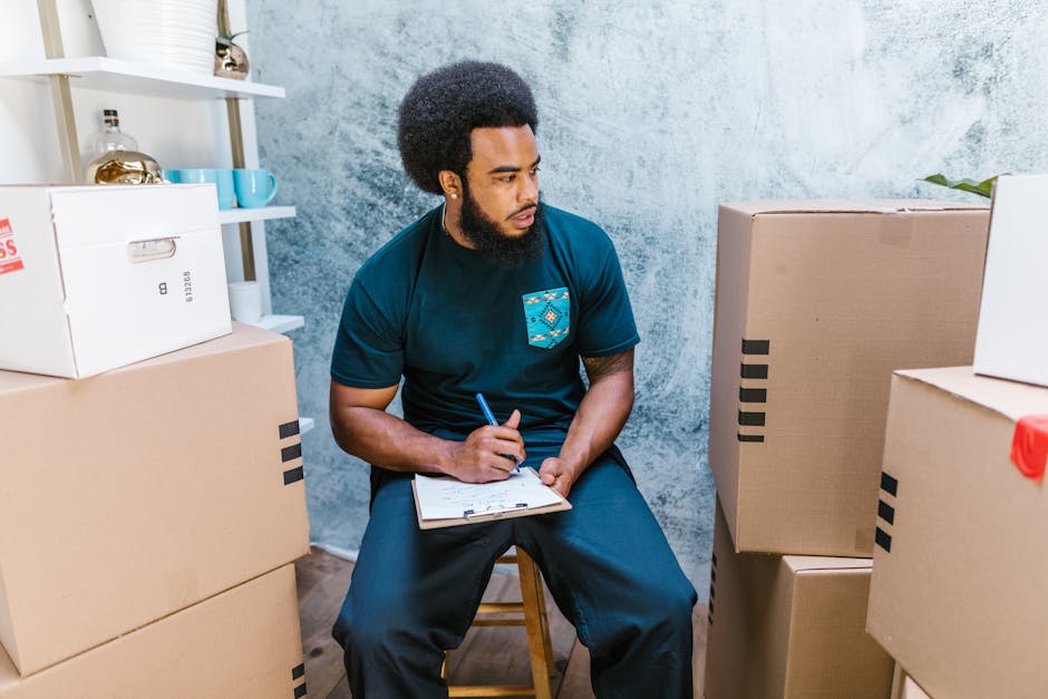 A woman with curly black hair, wearing a white t-shirt, blue jeans, and white sneakers, is seated among numerous large cardboard moving boxes inside a room with a light blue textured wall. The boxes, 